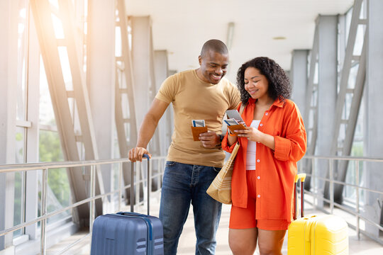 Young Black couple check travel information on a smartphone while holding passports and boarding passes beside luggage in a bright airport terminal before departure