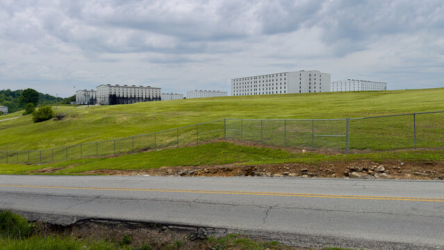 Rick houses, where bourbon is aged in oak barrels, overlook rolling green hills under heavy clouds; the left buildings show dark whiskey fungus staining, with a fence and road in front, Bardstown, Ken