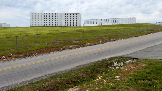 White rick houses, where bourbon is aged in oak barrels, crest a green hill behind a chain-link fence, with a cracked two-lane road and yellow center line in the foreground, Bardstown, Kentucky