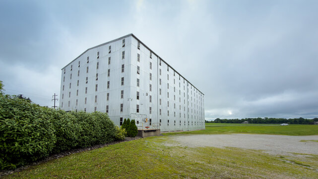 Corner of a tall metal-clad rick house marked 101 Bonded Warehouse, with a loading dock, hedge, gravel lot and rows of small windows under gray clouds, Bardstown, Kentucky