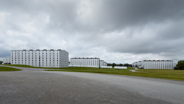 Several white rick houses, where bourbon whiskey is aged in oak barrels, stretch across a rural distillery campus, with a gravel lot in the foreground, parked trailers, and dark storm clouds above, Ba