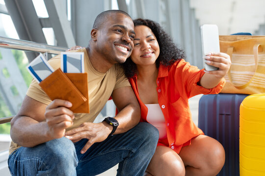 Smiling young Black couple taking smartphone selfie while sitting beside colorful luggage at airport, holding passports and boarding passes before their vacation flight
