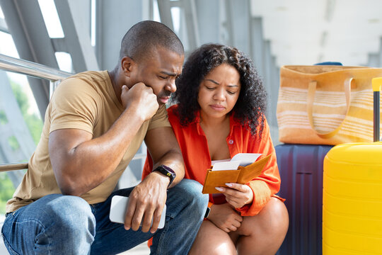 Concerned young Black couple sitting beside luggage at airport while checking passports, boarding passes and travel details, appearing worried about a problem before departure