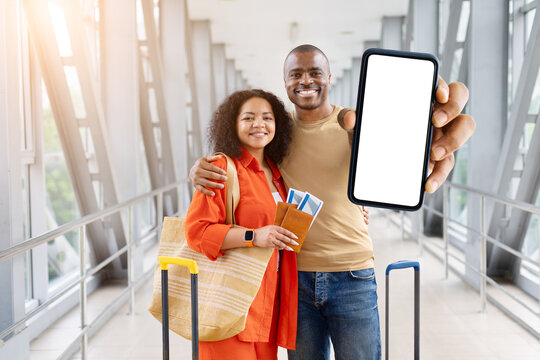 Smiling black couple holding passports at airport while showing blank smartphone screen to camera, mockup template with luggage nearby