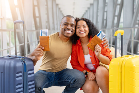 Smiling couple posing closely together holding up passports and tickets at airport terminal, crouching between colorful suitcases in a bright airport terminal before departure
