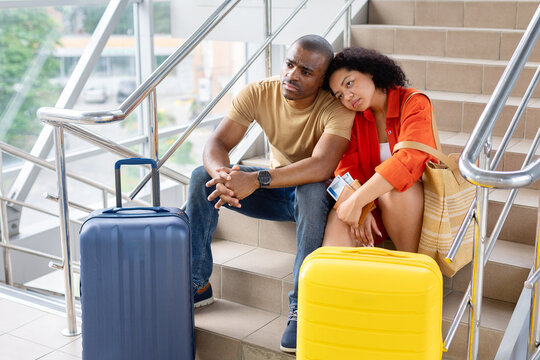 Young Black couple sitting tired and disappointed on airport stairs beside their luggage, waiting with passports and boarding passes during a stressful vacation journey