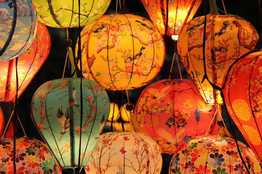 Horizontal composition of glowing traditional silk lanterns with oriental bird and cherry blossom patterns hanging at a night market in Hoi An, Vietnam.