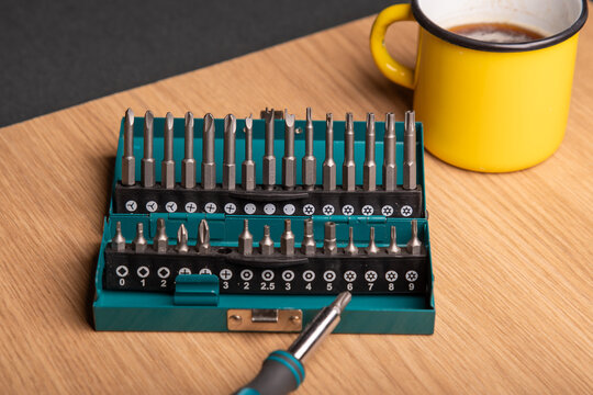 Handle of a screwdriver in front of a toolbox with tooltops for different screws. A yellow mug with coffee in the backgrond.