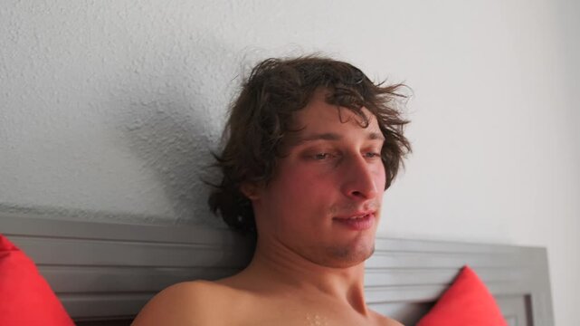 Close-up of a handsome young man with a dreamy look, lying in bed with red pillows after waking up. He gazes at his smartphone with a relaxed and sensual mood. Bright bedroom interior.