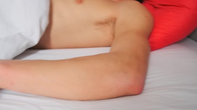 Relaxed young man sleeping shirtless under a white blanket, lying on red pillow in cozy bedroom. Peaceful rest, comfort and morning relaxation at home.