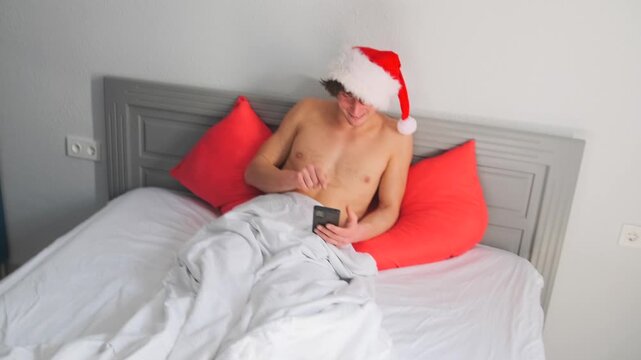 shirtless white man wearing santa hat reclining in bed while scrolling smartphone, red pillows, rumpled white sheets, soft morning light, relaxed smile, cozy holiday solitude, casual bedroom scene