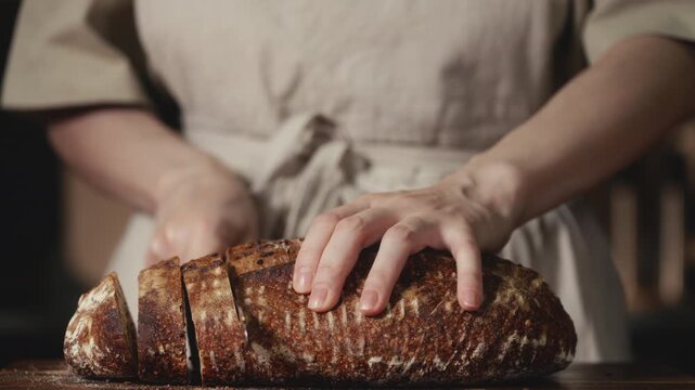 Close-up of baker in rustic linen apron slicing freshly baked artisan sourdough bread with knife on wooden cutting board. Homemade crusty loaf for breakfast, bakery promos, and healthy food concepts.