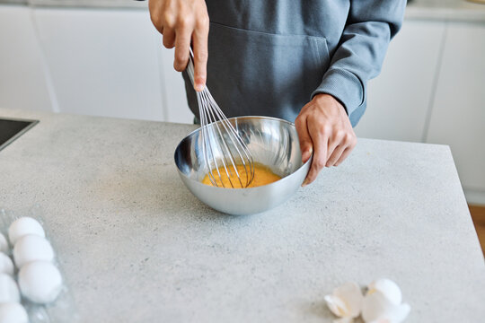 A teen cracks an egg into a stainless steel mixing bowl on a kitchen countertop, preparing to whisk for baking or breakfast. Visible hands, a whisk and an egg carton suggest home cooking, learning and