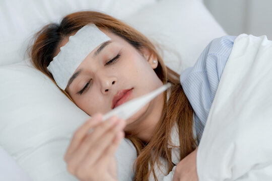 Sick young Asian woman lying in bed with a cooling patch on her forehead, looking at a thermometer while resting under a blanket at home, fever, illness, temperature check, recovery