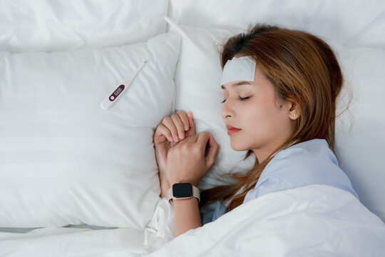 Sick young Asian woman lying in bed with a cooling patch on her forehead, looking at a thermometer while resting under a blanket at home, fever, illness, temperature check, recovery