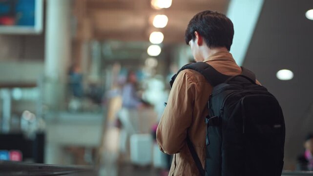 Rear view of young Asian man with backpack descending escalator inside modern airport terminal, male traveler moving through departure area with luggage, air travel, transportation, journey