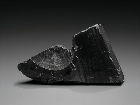 Sharp, jagged piece of black obsidian volcanic glass with a smooth, concave conchoidal fracture surface, resting on a neutral grey background