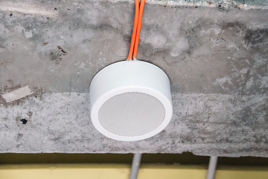 White circular speaker mounted on a raw concrete ceiling, showing exposed orange electrical wiring and industrial design