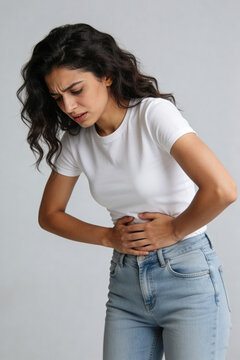 Woman holding her abdomen and looking down against plain background. Abdominal pain and digestive health concern. Healthcare, wellness and body care concept. Human health awareness.