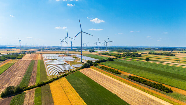 Solar installation and wind generators in rural area. Rows of photovoltaic modules occupy land next to tall vertical towers under bright sky.