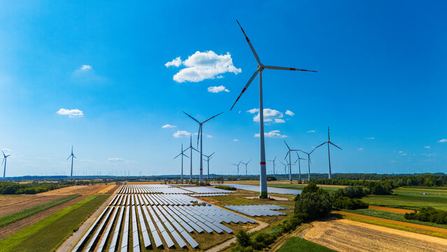 Solar panel layout and wind towers under cloudy sky. Scattered cumulus clouds drift above energy installations deployed on cultivated earth.