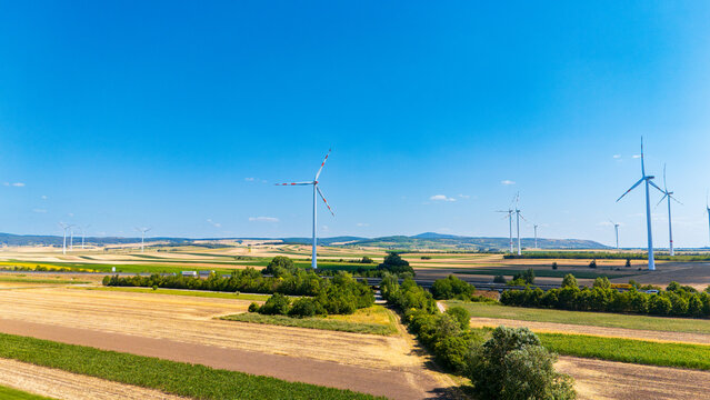 Wind generators above highway and agricultural terrain. A transportation route cuts through segmented crop fields beneath a clear blue atmosphere.