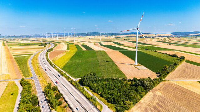 Wind turbine standing near highway and agricultural fields. Aerial view of a tall wind turbine beside a busy highway with agricultural fields under a clear blue sky.