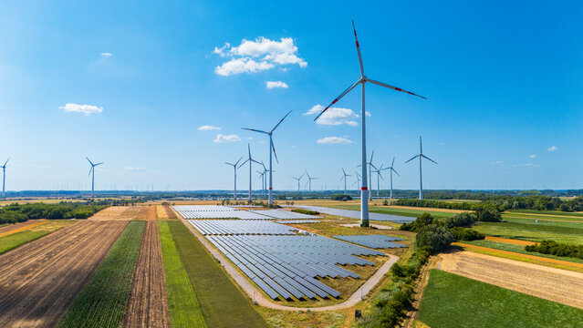 Solar array and wind towers in rural agricultural setting. Photovoltaic rectangles cover ground adjacent to towering vertical structures under daylight.