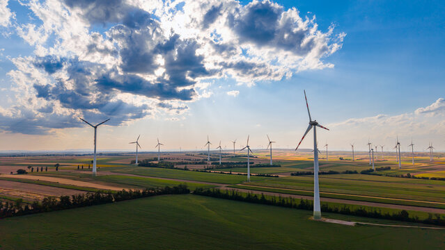 Wind turbines on agricultural fields under cloudy sky. Aerial view shows multiple white wind generators installed across green and brown rural farmland during daytime.