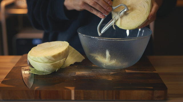 Close-up of woman's hands shredding fresh cabbage with a peeler into a glass bowl on a wooden board on a table. Cooking and healthy food preparation concept in a cozy warm home kitchen.