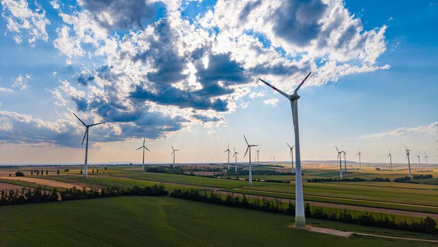 Wind turbine installation in rural agricultural territory. Overhead perspective captures numerous energy towers standing among cultivated patches beneath a bright sky.