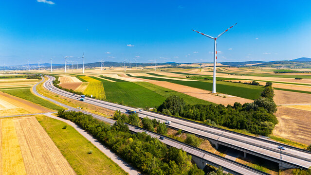 Highway passing through agricultural land with wind turbines. Aerial view of a highway running through colorful farm fields with wind turbines under a bright blue sky.
