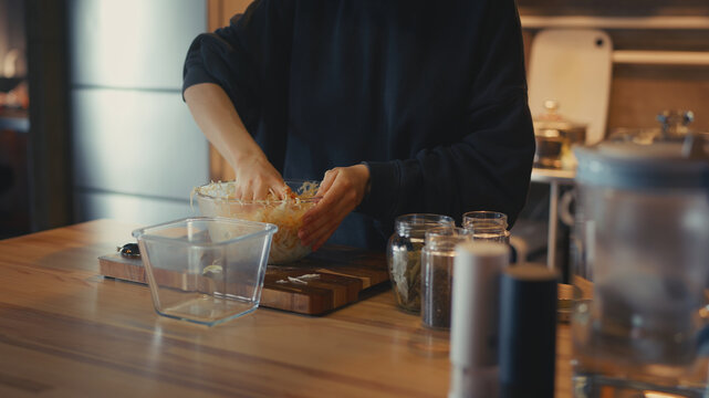 Woman hand mixing shredded cabbage and carrots with spices in a glass bowl. Preparing homemade fermented sauerkraut on a wooden table in a cozy kitchen. Close-up process of salting vegetables.