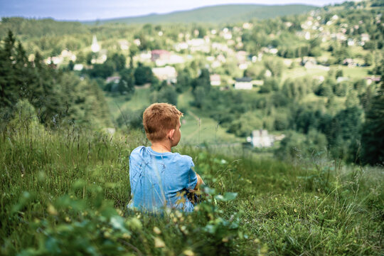 Boy sitting alone on mountain hillside overlooking countryside, childhood, solitude, reflection, nature and outdoor adventure concept
