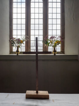 A simple wooden cross stands before a large, mullioned window in a dimly lit room, with vases of wildflowers on either side. The scene evokes a sense of quiet contemplation and natural beauty.
