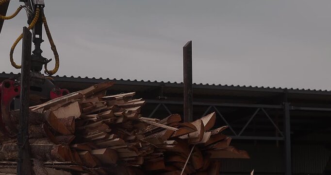 Hydraulic grapple lifts bundled logs and timber boards at an outdoor lumber processing yard with industrial shed and stacked wood materials