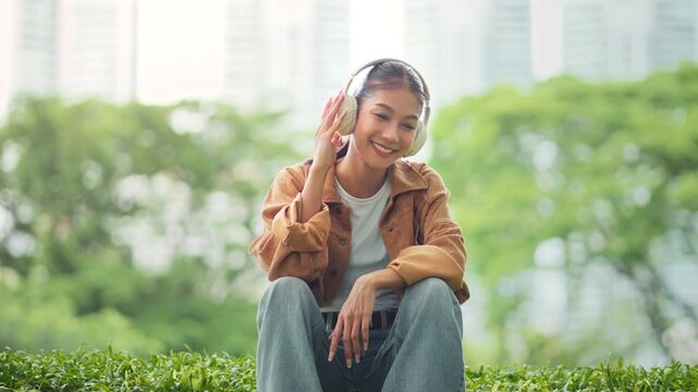 Happy young Asian woman wearing wireless headphones and gently moving to music while sitting on grass in urban park, relaxed female enjoying rhythm outdoors