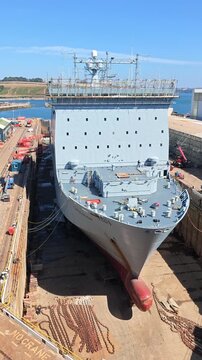 Falmouth Cornwall England UK.  21.08.2026. Video. A grey British warship sits in a dry dock for repairs with industrial cranes and shipyard buildings visible in the background on a sunny day