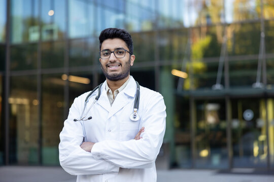 Young indian male doctor in white lab coat with stethoscope, standing outdoors with arms crossed and smiling confidently in front of a modern medical building