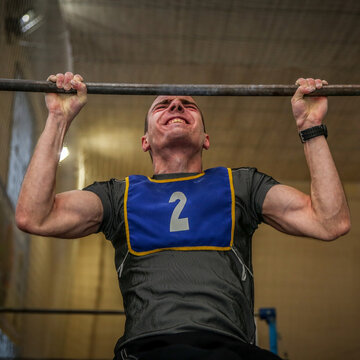 Strong athletic man doing pull-ups on horizontal bar during fitness crossfit workout or competition. Caucasian sportsman training chin-ups with intense facial expression in indoor gym