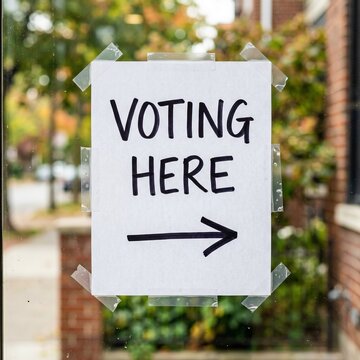 A White Sign Stating Voting Here Being On Hold With Adhesive Tape Applied On A Clear Glass Window.