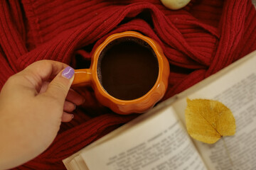 Obraz premium An orange mug of coffee resting on a red knitted sweater beside an open book and a yellow autumn leaf, with soft film grain, high quality photo