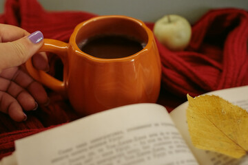 Fototapeta premium An orange mug of coffee resting on a red knitted sweater beside an open book and a yellow autumn leaf, with soft film grain, high quality photo