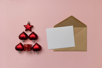 Flat lay of kraft envelope with blank card and red ornaments arranged as a Christmas tree on smooth pink background. © tatiana