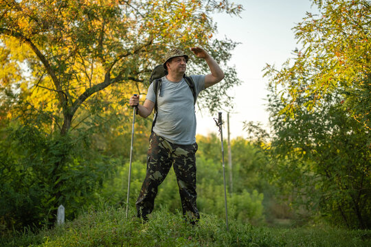 Hiker pauses on green woodland trail, gripping trekking poles and looking ahead with alert concentration. Backpack and hiking clothes fit active outdoor travel beneath sunlit autumn foliage.