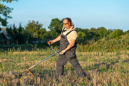 Man uses grass trimmer across dry field, standing among cut stalks. Equipped with hearing protection and work overalls, landscaper performs outdoor upkeep with calm, focused expression.