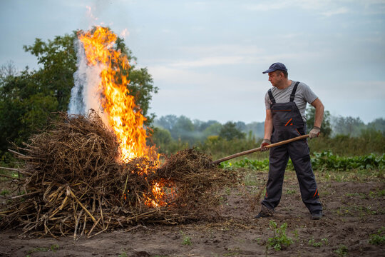 Adult man controls brush fire on muddy farmland. Wearing gloves and work overalls, he prods dry plant debris with pole while watching bright flames, creating tense scene of careful land clearing.