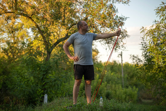 Man pauses with resistance band on green hillside, using one hand for support and one at his hip. Serious adult waits in calm evening light, framed by trees and natural woodland in standing pose.