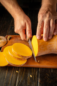 Hands prepare butternut squash by slicing it on cutting board. Sharp knife separates thick rounds from vegetable, and close view gives this kitchen task focused, practical feeling.