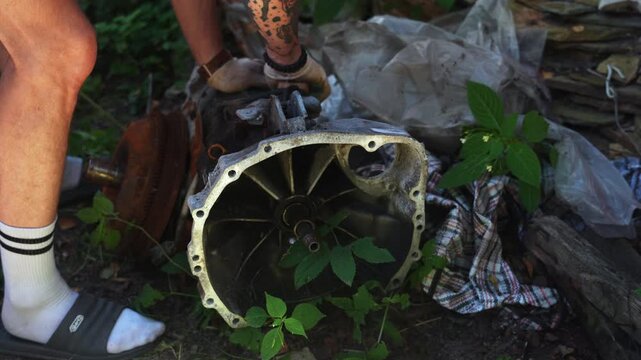 A man finds a car transmission in the backyard, examines it, and wipes off the dust